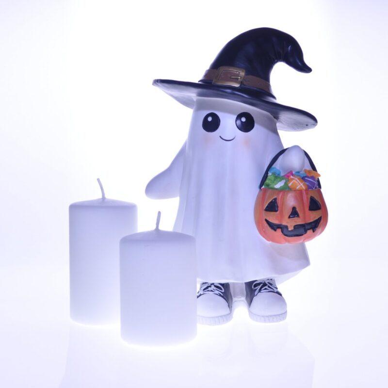 Zestaw dwóch białych świec o gładkiej powierzchni oraz ceramiczna figurka ducha w kapeluszu czarownicy i z dyniowym koszykiem. Kompozycja ustawiona na białym tle. Idealna dekoracja do wnętrz lub jako prezent.
